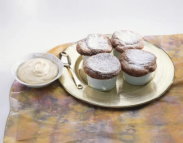 Four sugar-sprinkled mini souffles in cups on metal plate, spoon, bowl of cream, table cloth, close up