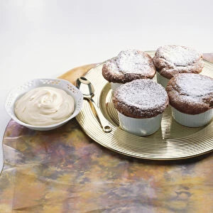 Four sugar-sprinkled mini souffles in cups on metal plate, spoon, bowl of cream, table cloth, close up