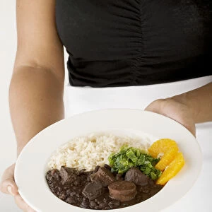 Women holding plate of feijoada, a bean and meat stew, served with rice, orange slices and kale